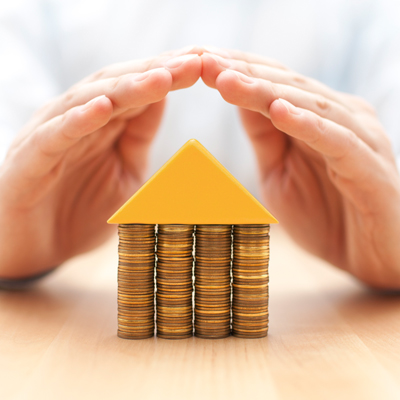 hands covering a house made by 4 stacks of coins and a yellow triangular cylinder
